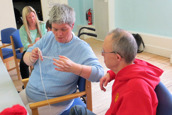 photo: A woman in blue is making something with white wool. She is showing a man who is sitting next to her what she is doing.
