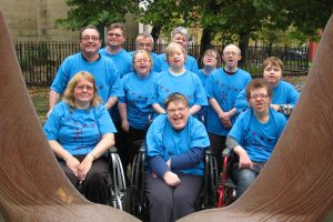 We can see a about a dozen people all in blue t-shirts with the DIY logo on. They are gathered together to have their photo taken and are smiling at the camera