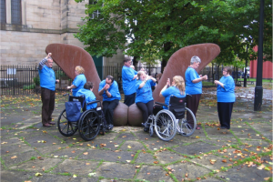 We can see people outside gathered round a large metal sculpture of a sycamore seed. The people are all wearing black trousers and bright blue t-shirts. They are talking to each other in small groups. Two of the people are in wheelchairs