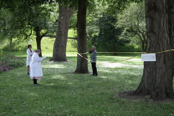 We can see David dressed as a patient in an institution. He is in an area on his own kept apart with yellow tape. We can also see Anna on the right dressed as a Doctor. She has a clip board and is looking at David sternly.