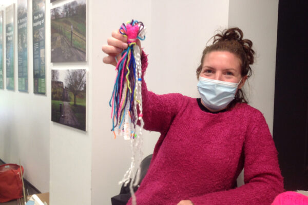 photo of DIY staff member Mary with a facemask on. She looks happy and is holding up her colourful tassle which contains written down her thoughts about lockdown.