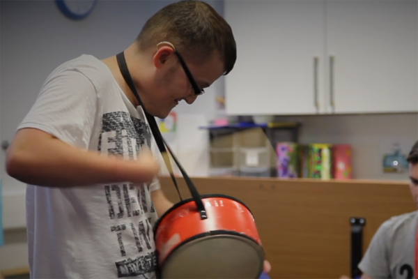 photo of student playing a drum. He is wearing glasses and a hearing aid. The drum is bright red and hangs round his neck. His hand is moving so fast it is just a blur!