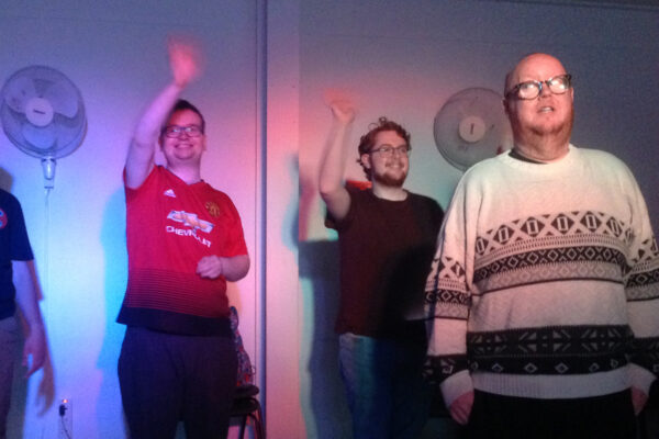 photo: 3 people are standing looking forward. They are brightly lit and 2 are holding a hand up waving.