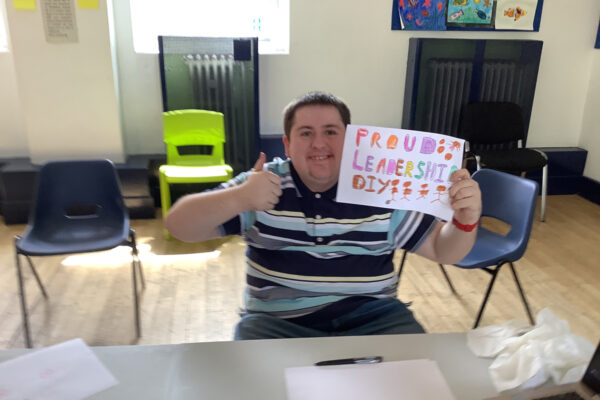 photo of a young many in a stripey shirt. He is smiling and holding up a poster which says 'Proud, Leadership, DIY'. It is very colourful and has some people drawn at the end. He is also showing us the thumbs up sign.