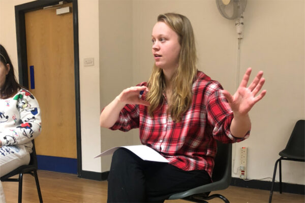 photo of a young woman sitting in a room. She is sitting on a black plastic chair and is wearing a red checked shirt. There is a piece of white paper on her knee, both her arms are raised; her right arm is bent with the hand facing down, her left hand is held up witht the palm facing us. She is looking to her right where we can just see another person also sitting on a black plastic chair.