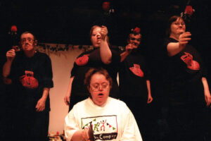 photo: Actors are on a stage. There is a person at the front in a white t-shirt sitting down. Behind her are 4 people dressed all in black but with a red symbol on their chests. These people are all holding up red roses.