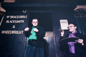 Two men are standing infront of a wooden building which is painted dark blue. There is neat white writing on the building 'Wm. Bracegirdle Blacksmith & Wheelwright'. We can also see a white poster with the word 'Notice' at the top. Infront of a doorway is a man dressed in black except for a green top. He is staring at the camera and has his hands infront of his chest in loose fists. To his right is another man who looks like an artist as he has a pain brush and large palet. He is looking at the 1st man.