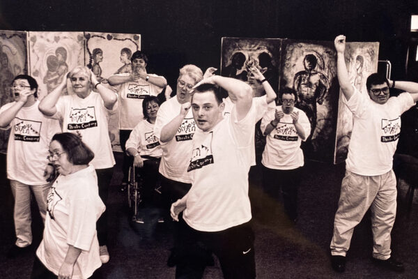 We can see a large group of people standing and one person in a wheelchair. The are all doing something different with their arms and look like they are doing action poses. They are all wiearing white t-shirts with the same logo on the front. A young man has one hand on their head, an older woman has her hands over her ears, a man further back has one are high in the air!
