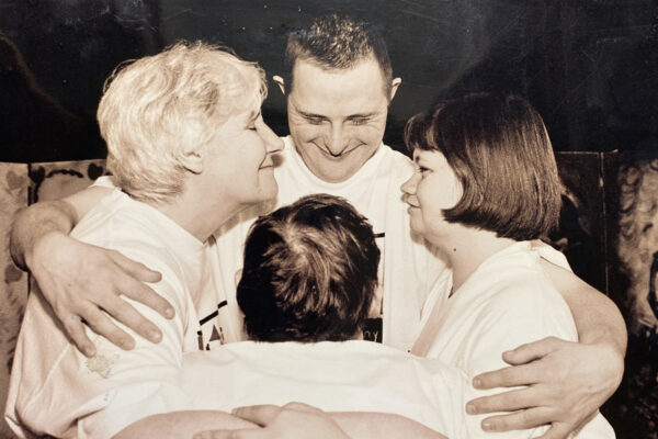 We can see 4 people facing each other in a big hug. They have their eyes shut and are smiling. They are wearing white t-shirts.