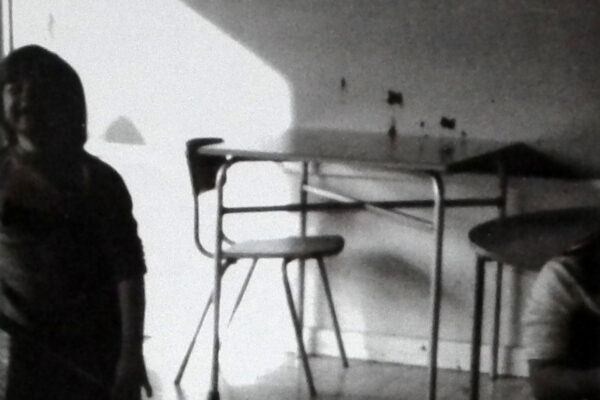 a black and white grainy image. There is a panel of light on the left, probably from a large window. There is a figure in the foreground on the left. The light is behind them so it is difficult to see but it looks like a child. Behind we can see a small dining table and 2 modern dining chairs.