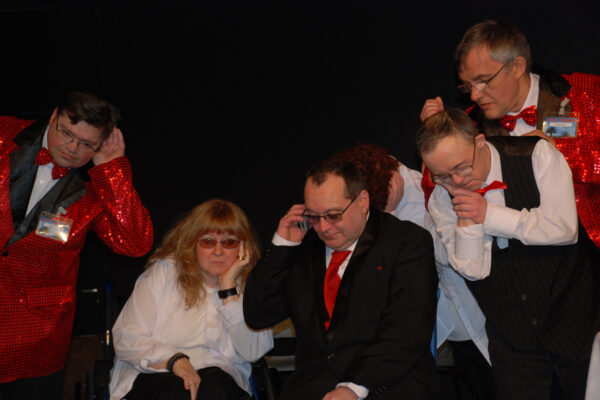 There are 5 people. In the centre are a woman and man, the man is holding a phone to his ear. Everyone else is leaning in listening. The people are dressed formally in suits and ties, although the suits of two of the men are unusual being bright red sparkly.