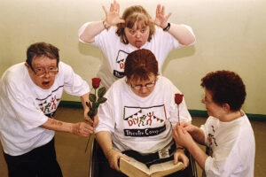 close up photo of 4 people in white t-shirts with DIY Theatre's logo on them. There is a woman in the middle who is sitting looking down at a big book which is open on her knees. On her right is a man who is holding out a red rose to her. On her left is a woman kneeling and also holding out a red rose to her. Behind her is a woman who leaning over her. This woman is not being very nice! She is sticking out her tongue and holding her hands up to her head like antlers. The woman reading seems to notice none of this!