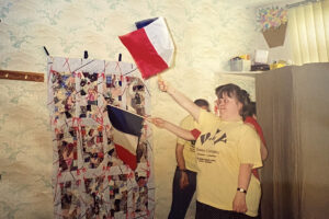 Photo of 3 people standing in front of each other. 2 of the people are waving oblong flags with a strip each of red, white and blue on them. There is a large white banner on the wall which has red and blue lines on it and 12 regular oblongs of what look like random collaged pieces of patterened material. We can see some are stiped red and white.