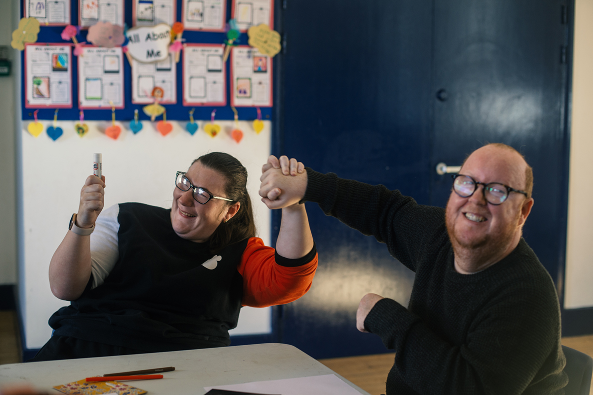 A young woman and a young man are sitting at a table. They look like they are messing about, having fun and laughing. They are holding each others hands and holding these up between them. The young woman is also holding up a pen or glue stick. On the desk in front of them are coloured pens and stickers alongside sheets of paper. Behind them is a large double door and a notice board with lots of pieces of paper on, coloured hearts and in the middle a piece which says 'All About Me'