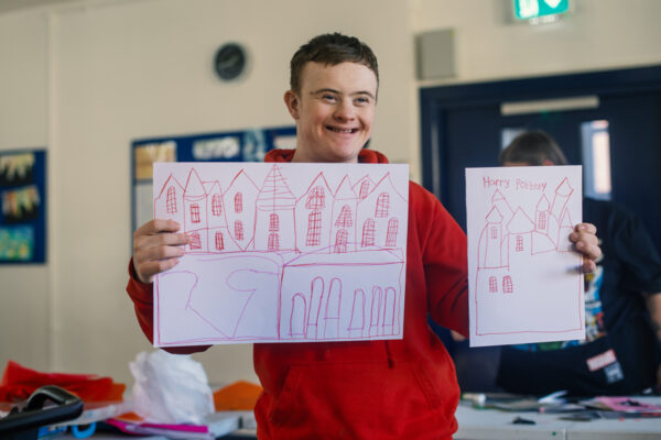 photo: a boy dressed in bright red is holding up 2 pieces of paper with drawings in red on them. Both drawings are of buildings. There is a large sheet in his right hand - this looks like a street with a row of houses with pointy roofs and windows with many small panes of glass. In his left hand he is holding up a smaller piece of paper with a drawing of a castle on with lots of turrets with pointy roofs. Again the windows are made up of lots of panes. Written at the top is 'Harry Pottery'. The boy has a big grin on his face and his eyes are all wrinkley in the corners because of this. It's a big smile!