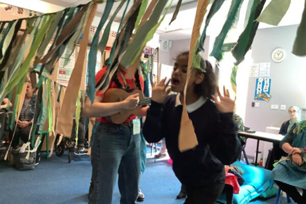 photo: a young person is moving through a string of hanging pieces of fabric. We just see through the fabric that someone is playing a guitar. There is a lot of light in the room