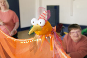 photo of a puppet bird. It is bright orange with big round eyes, it's wings are made up of transluscent orange fabric and have a row of small lights twinkling at the top. We can see two people behind the puppet who are operating it, one is in a wheelchair.