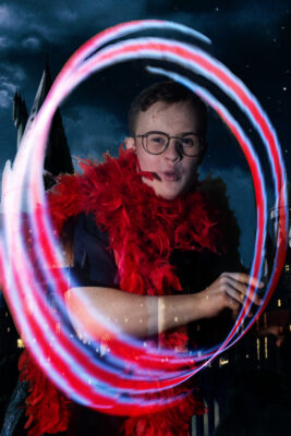 photo: a very young man is looking at us through a large oval formed of red white and blue lines of light. He looks like he might have just made this happen - he looks excited and his lips are pursed as if he is saying 'ooh' He has short dark hair and is wearing glasses with slim frames. Around his neck is a red feather boa.