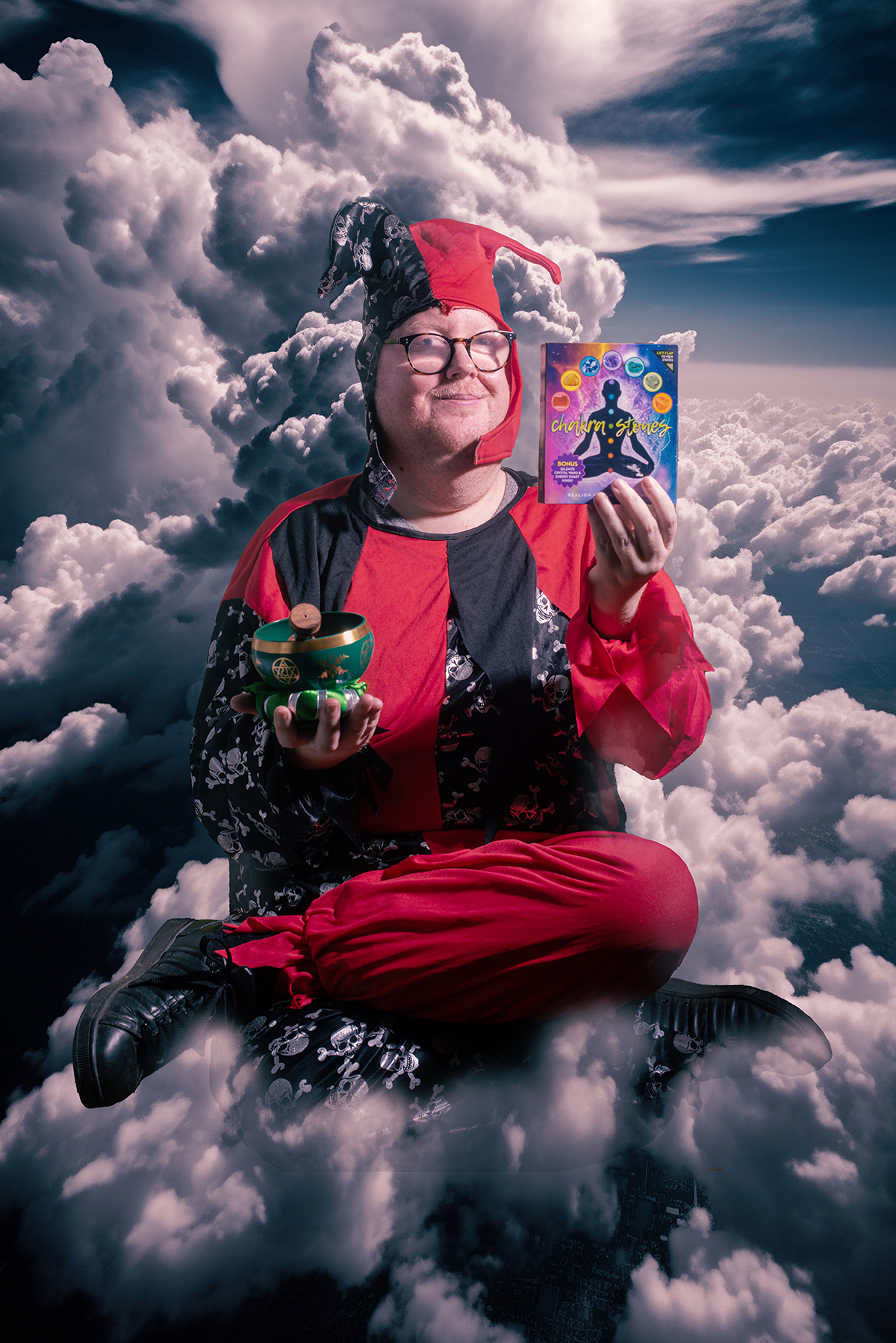 photo: a man dressed in a red and black striped jesters costume is sitting cross legged ontop of clouds. If you look closely you can see that there are skull and crossbone images on the black parts. The man is looking at us and holding things in his hands. In one hand he has a dark green bowl with gold symbols on it. This is resting ontop of a small green cushion on the palm of his right hand. In his left hand he is holding up a book sized box. On this we can see 'Chakra Stones' and the silouette of someone sitting with crossed legs. It has lots of different coloured circles on it.