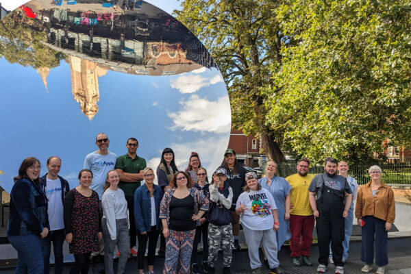 photo: its a sunny day in an urban space. A large group of people are standing together to have their photo taken. They are casually dressed and smiling. Behind them is a very large round mirror - it is over twice the hight of the tallest person. It has an upside down reflection in it showing lots of blue sky and what may be a church tower in pale stone.