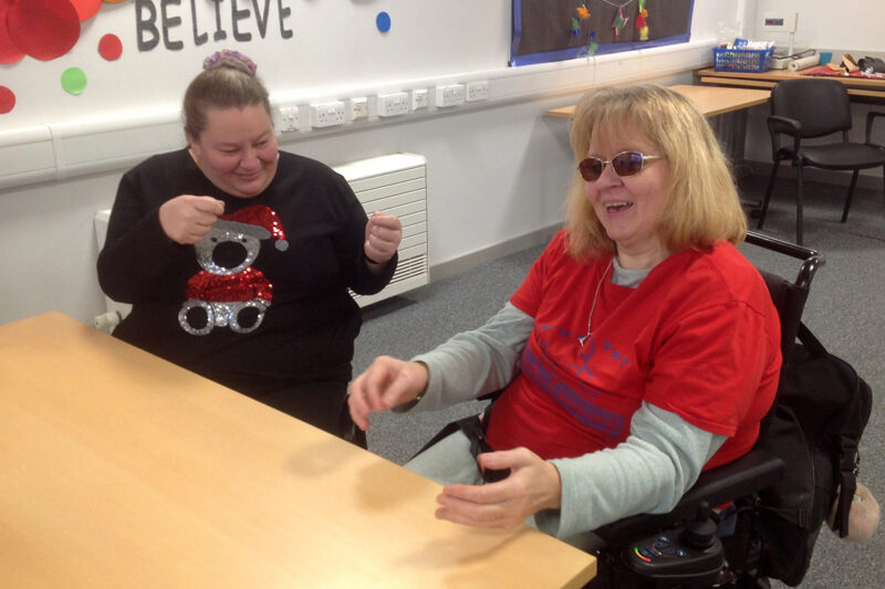 photo: 2 women are sitting beside a table. One is in a red t-shirt with the DIY logo on. She is talking and doing something with her hands - as if there was something there that we cant see. The other woman looks like she is holding something in her hands and looking at it - but again there isnt anything there.