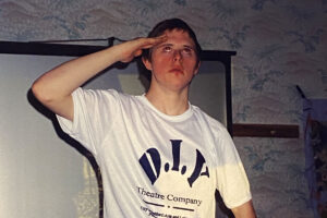 photo: a young man is saluting with his hand to his head and looking up. He is wearing a white t-shirt with "D.I.Y." in big black letters