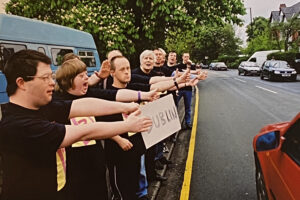 photo: the right half of the photo is of a road going off into the distance. On the left and at the edge of the road there is a crowd of people holding their thumbs out to try and hitch a ride. One is holding a piece of cardboard with 'DUBLIN' written on it.