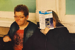 photo: close up of 2 people infront of an old interior wall. The person on the left is looking slightly off to the right and is holding her lips close together. we can't see the other persons face as they are holding up a booklet to look closely at it.