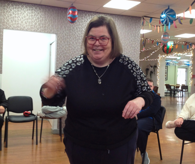 A smiling woman wearing glasses and a black top with patterned sleeves raises her arm during a DIY Theatre session. Chairs and colourful decorations are visible in the background.