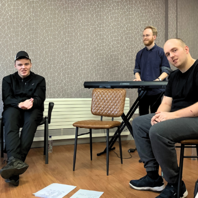 Three men in a creative session, seated and standing in front of a geometric-patterned wall. One man, wearing a black cap and jacket, sits on the left, smiling with his hands clasped. Another man, in a black T-shirt and grey trousers, sits on the right, leaning forward and smiling. A third man, wearing glasses and a navy shirt, stands behind them at a Yamaha keyboard, looking engaged. Two sheets of paper with notes are on the wooden floor in the foreground.