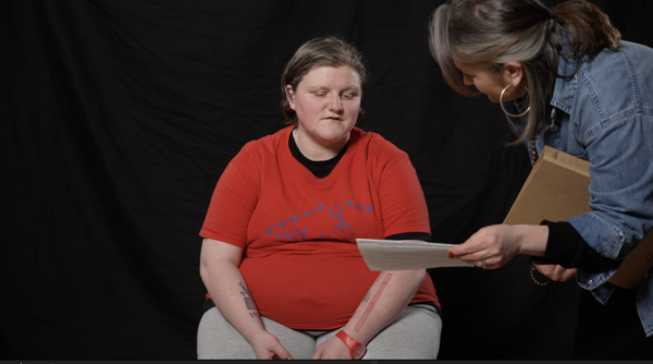 Charlotte is wearing a red DIY Theatre Company T-shirt sits against a black backdrop, looking down at a piece of paper. Hilary has a grey-streaked ponytail, wearing a denim jacket, leans in while holding documents and offering guidance. The image captures a moment of focus and collaboration.