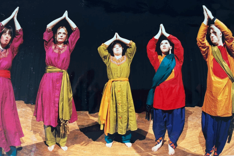 Six performers stand barefoot on a stage with their arms raised above their heads, palms pressed together in a prayer-like pose. They wear vibrant, coordinated costumes featuring tunics in colours like magenta, green, red, and orange, paired with loose trousers and contrasting sashes. The black stage backdrop highlights the rich textures and colours of their clothing. The image captures a powerful and unified moment in a performance by an inclusive dance or theatre company, evoking themes of focus, balance, and collective expression.