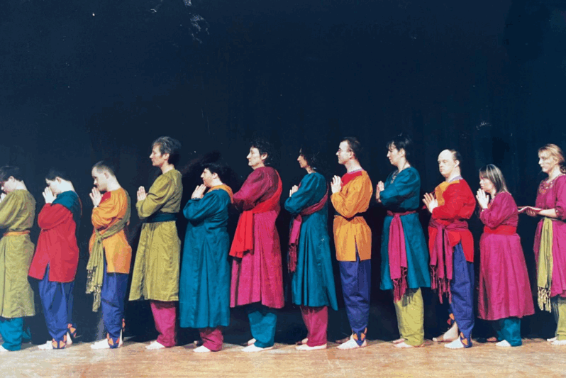 A diverse group of performers stand in a single line on stage, all facing the same direction with their hands pressed together in a prayer-like gesture. They wear colourful, coordinated costumes featuring tunics in shades of magenta, teal, mustard, and orange, with matching sashes and loose trousers. The barefoot performers, including people with and without learning disabilities, are captured in a moment of calm focus against a dark theatre backdrop. The image conveys unity, discipline, and inclusivity, likely from a performance by an integrated dance or theatre company.