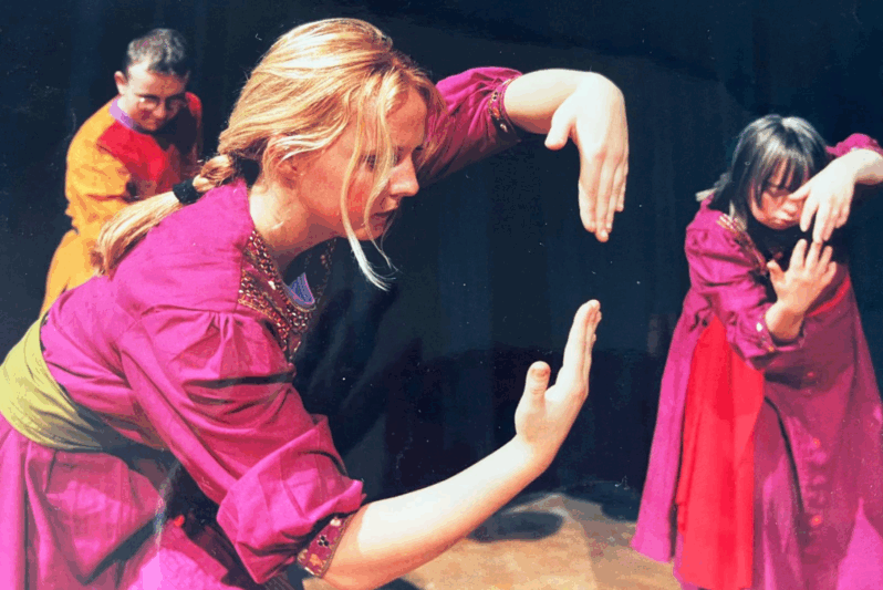 Three performers are captured mid-movement during a stage performance, each deeply focused on an expressive gesture involving their hands. In the foreground, a blonde woman in a magenta costume with a green sash curves her hands in a circular shape, creating a frame in the air. To her right, a young woman in a matching costume mirrors the motion with intense concentration. In the background, a performer in orange and red watches attentively. The scene is intimate and powerful, reflecting a moment of storytelling or emotional expression in an inclusive dance or theatre piece.