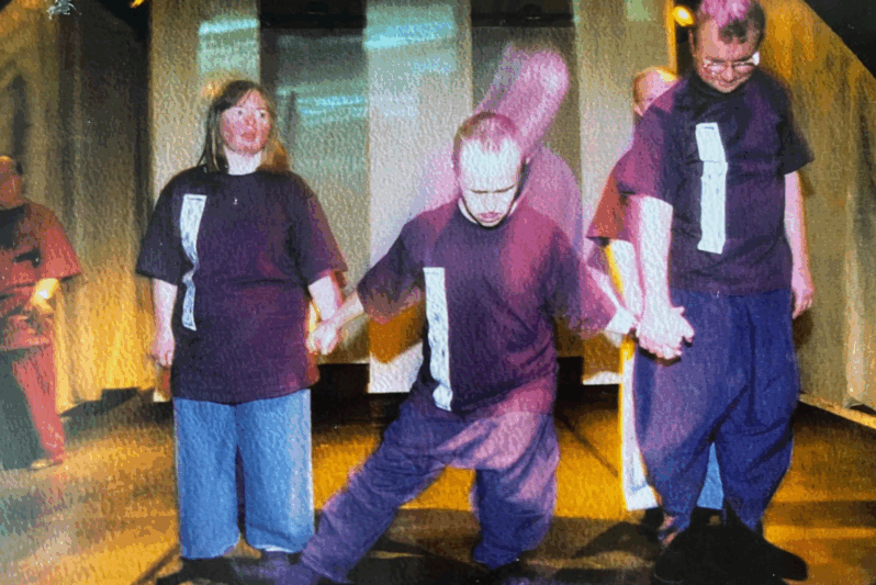 A group of performers stand on stage holding hands, wearing matching dark purple shirts with vertical silver patches and loose blue trousers. The central performer is in motion, dipping into a deep lunge while the others remain upright and connected. The lighting casts warm tones and shadows across the scene, creating a powerful, intimate atmosphere. The image captures a moment of synchronised movement and solidarity during an inclusive theatre or dance performance.