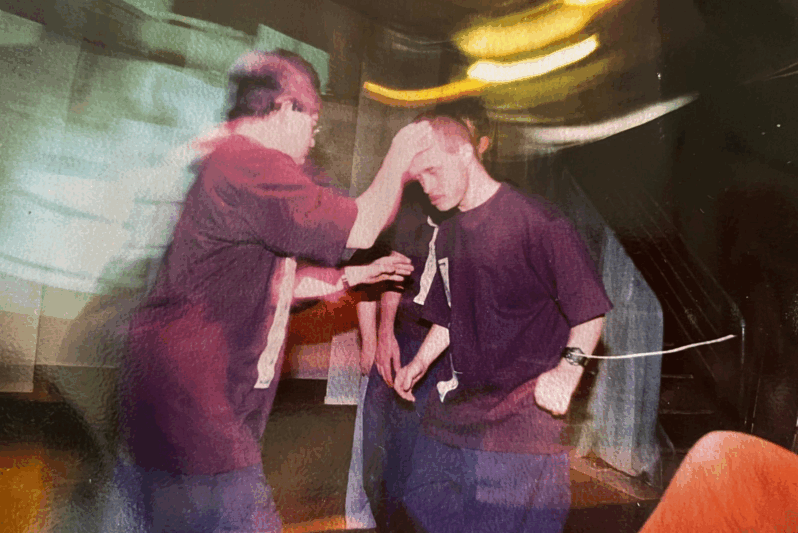 A motion-blurred image shows two performers in dark purple costumes engaging in a theatrical interaction. One performer reaches out gently to touch the forehead of another, who bows slightly with eyes closed, suggesting a scene of blessing, healing, or connection. The dim lighting and abstract movement trails evoke emotion and intensity, reflecting a meaningful moment in a devised performance by an inclusive arts company.
