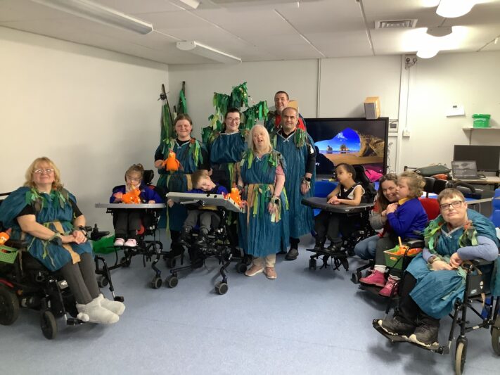 A group of adults and children, some using wheelchairs, pose together in a classroom setting. They are all wearing matching blue costumes decorated with green leaf-like fabric, possibly for a performance or themed activity. Some people are holding orange stuffed animals. The atmosphere is cheerful and inclusive, with a background showing a computer monitor, desks, and classroom materials.
