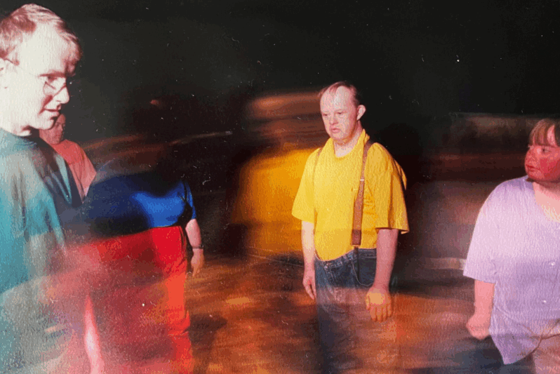A group of performers stand on a dimly lit stage, captured with motion blur that gives a sense of movement and atmosphere. The central figure, a man in a bright yellow shirt with suspenders, gazes ahead with a thoughtful expression. Other participants, wearing vibrant colours like purple, red, and teal, are partially blurred as they move through the space. The dark background and dynamic lighting create a dramatic, immersive feel, suggesting a rehearsal or performance by an inclusive theatre or dance company.