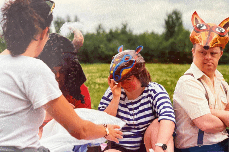A group of people are seated outdoors on a grassy field, participating in a creative arts session. Several individuals wear brightly coloured, handmade animal masks, including a fox, a ram, and a multicoloured creature with ears. A woman in a striped top adjusts her mask while another woman with her back to the camera, wearing a white T-shirt and sunglasses on her head, offers a white object—possibly part of the activity. The atmosphere is warm and communal, suggesting an inclusive arts workshop, likely facilitated by the Dodeka Dance Company. Trees and an overcast sky form the backdrop.