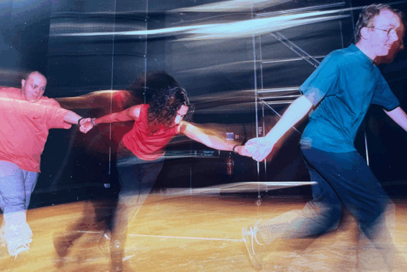 Three performers holding hands in a dynamic, motion-blurred moment on stage. The person on the left wears a red T-shirt and grey joggers, the person in the middle is Jude Bird and she wears a sleeveless red top, and the person on the right wears a teal shirt and glasses. They appear to be mid-run or dance in a theatre setting with scaffolding and stage lighting in the background.