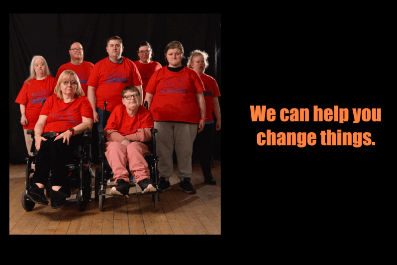 A group of eight members of DIY Theatre Company stand and sit together in a performance space. Two women in wheelchairs are at the front, with six others standing behind them. They all wear matching red DIY Theatre t-shirts and look directly at the camera against a black backdrop. To the right of the image, bold orange text reads: “We can help you change things."